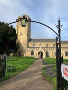 Image shows a a view through a metal arch and up a pathway to the full facade of a traditional english stonebuilt church with a tower