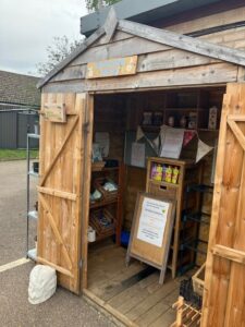 Image shows a small wooden structure with double doors open. Inside is a variety of shelving on which is displayed longlife foodstuffs.